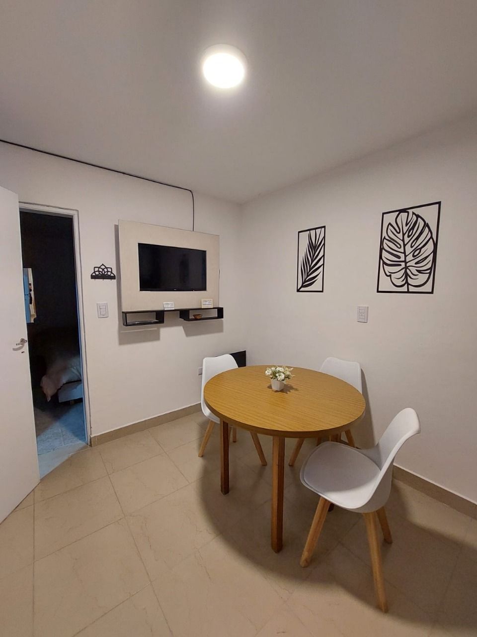 Pequeño comedor con mesa redonda de madera, sillas blancas, TV y arte decorativo de pared.
