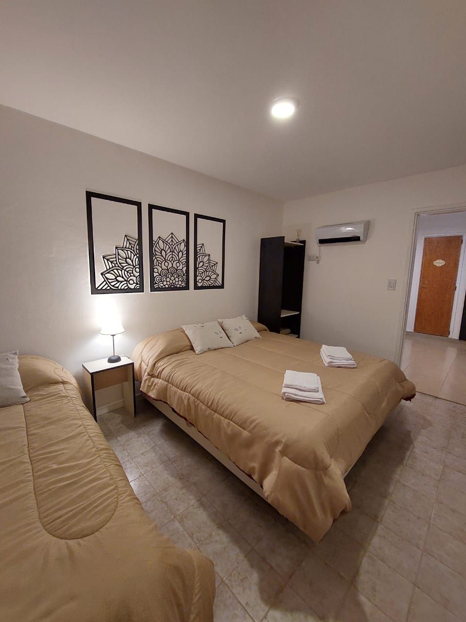 Dormitorio con una cama doble, una cama individual y obras de arte. Paredes blancas, ropa de cama marrón y suelo de baldosas.