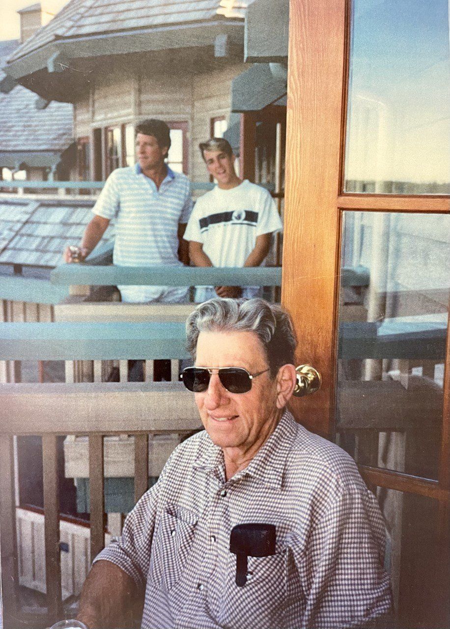 Man in sunglasses seated on a deck, two men in background. Wooden structure, sunny day.