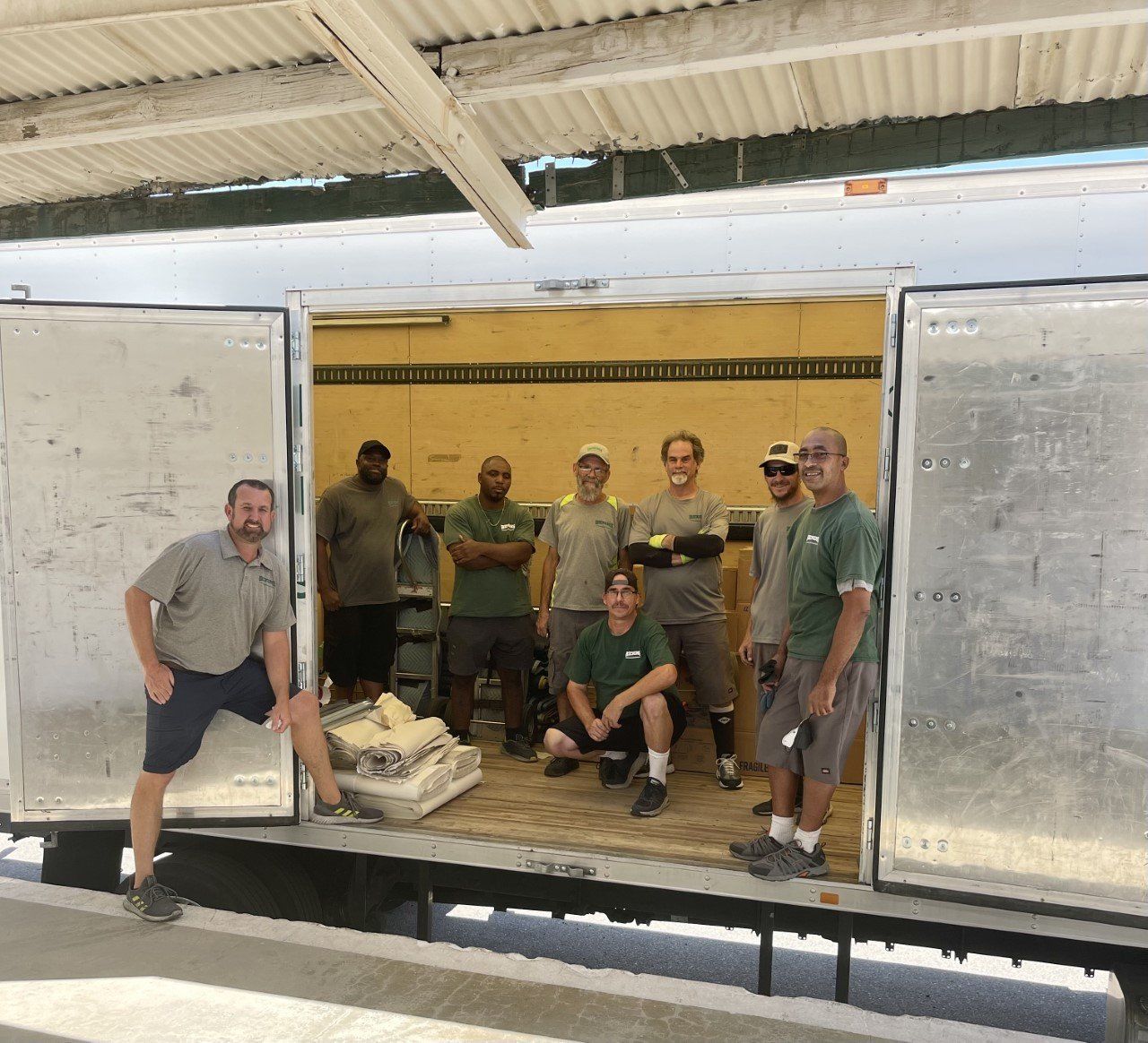 Group of men inside a truck with open doors, posing. Some are standing, and one is kneeling near a large object.