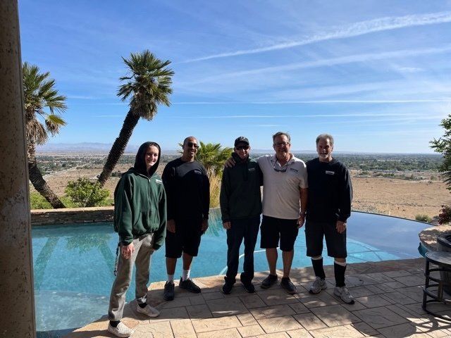 Five people standing by an infinity pool, desert landscape in background. Sunny, blue sky.