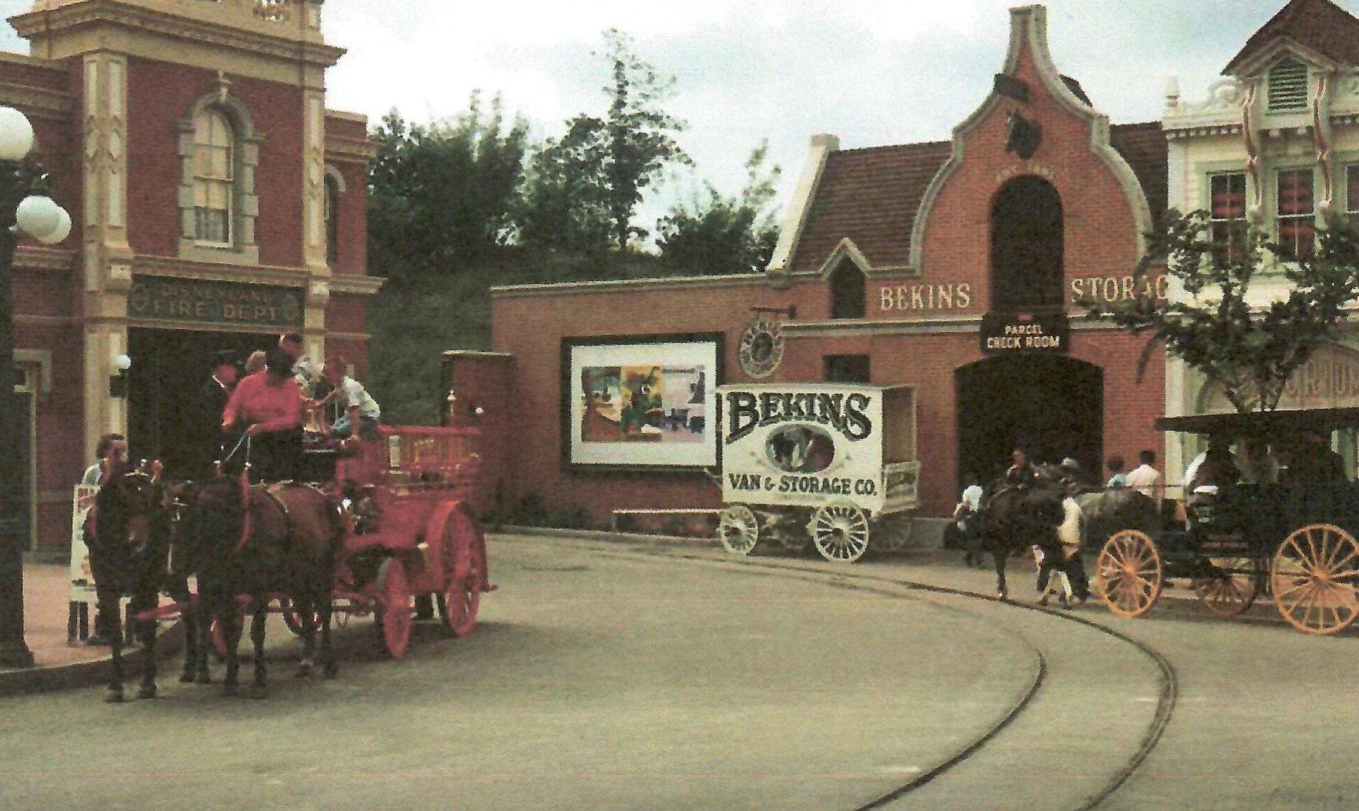 Horses pulling a red fire engine on a street with old-fashioned buildings. A sign says “Beniks”.