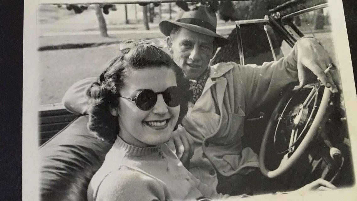 Woman in sunglasses smiles in a vintage car. Man in hat is in the back.