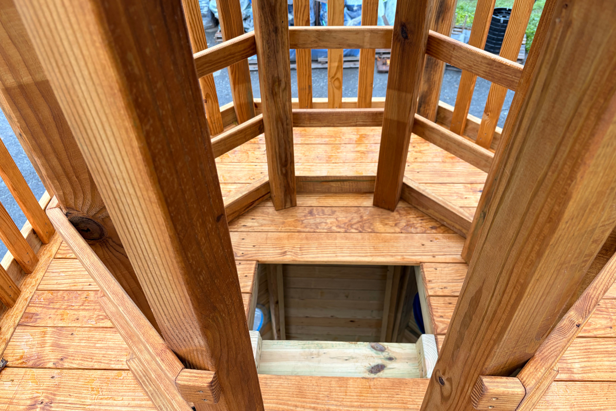 View of the upper observation deck on a custom wooden lighthouse playground, showing the ladder opening, natural wood flooring, support posts, and surrounding wooden railings.