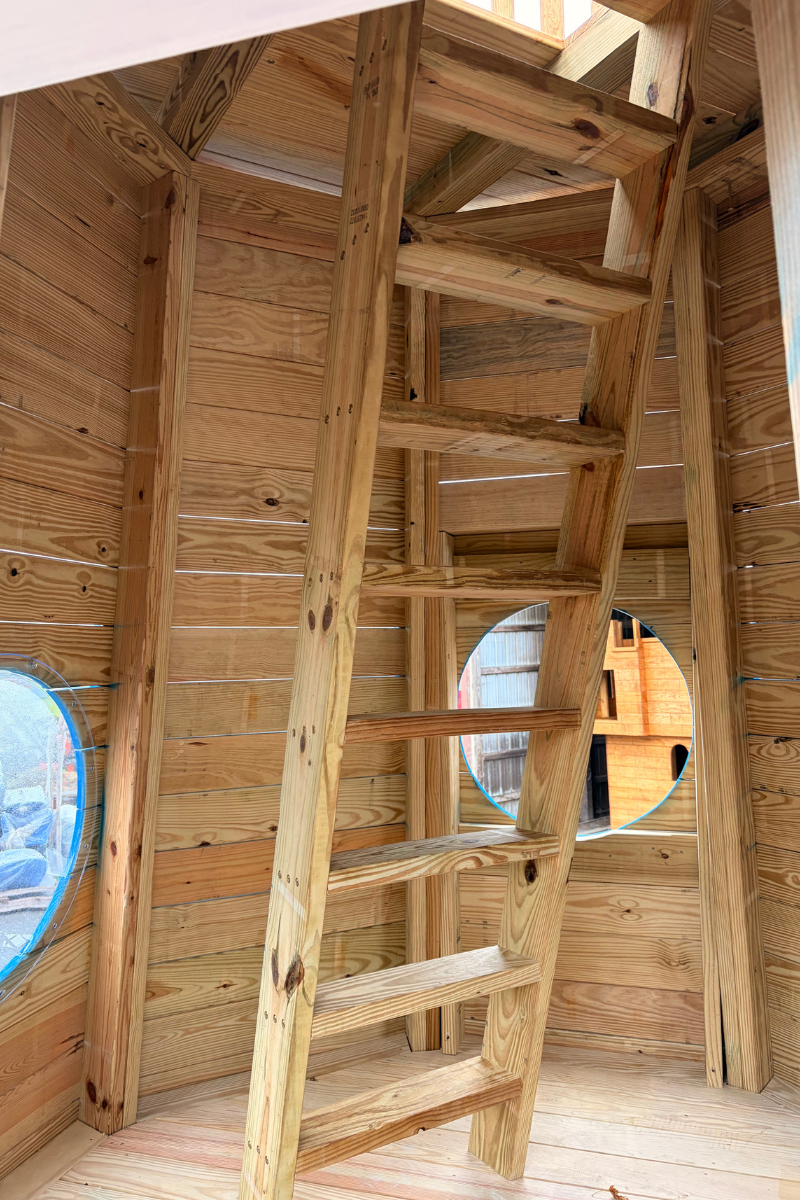 Interior of an Amish-built wooden lighthouse playground featuring a climbing ladder leading to the upper observation deck, natural wood walls, and round windows.