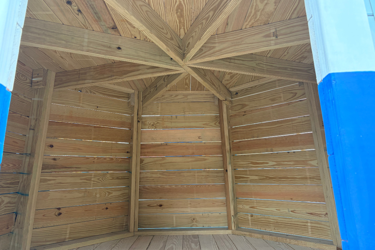 Ground-level entrance to an Amish-built wooden lighthouse playground, featuring an open playhouse interior with natural wood plank walls, flooring, and exposed ceiling beams.