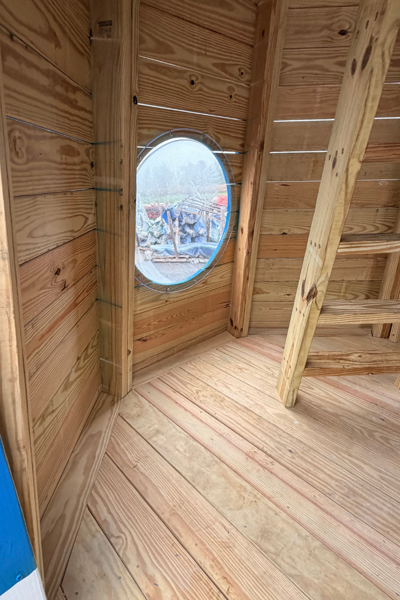 Interior of an Amish-built wooden lighthouse playground featuring a clear bubble window, natural wood flooring, and a climbing ladder leading to the lookout deck.