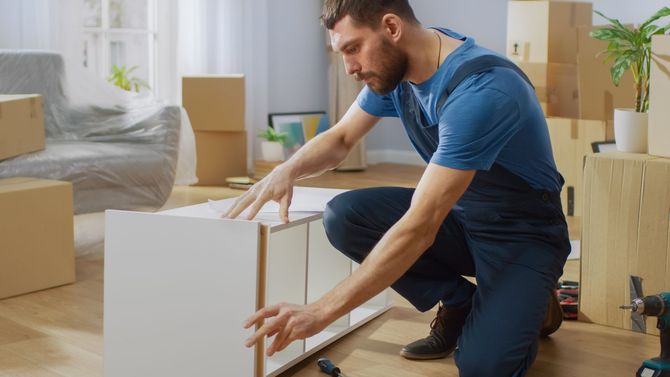 A man is kneeling down in a living room while assembling a cabinet.