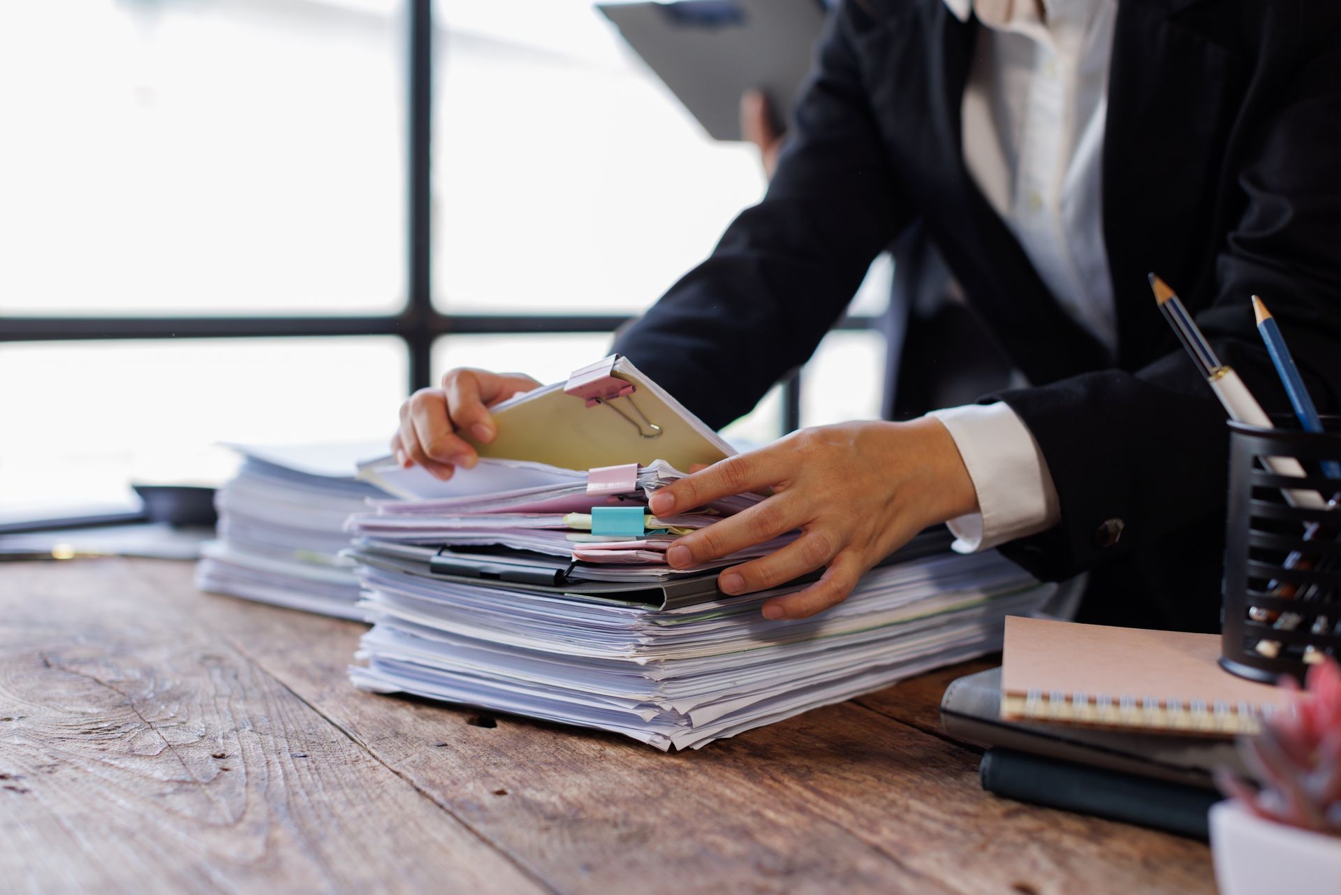 Person in a blazer sorting through a large stack of documents at a wooden desk, near a window.