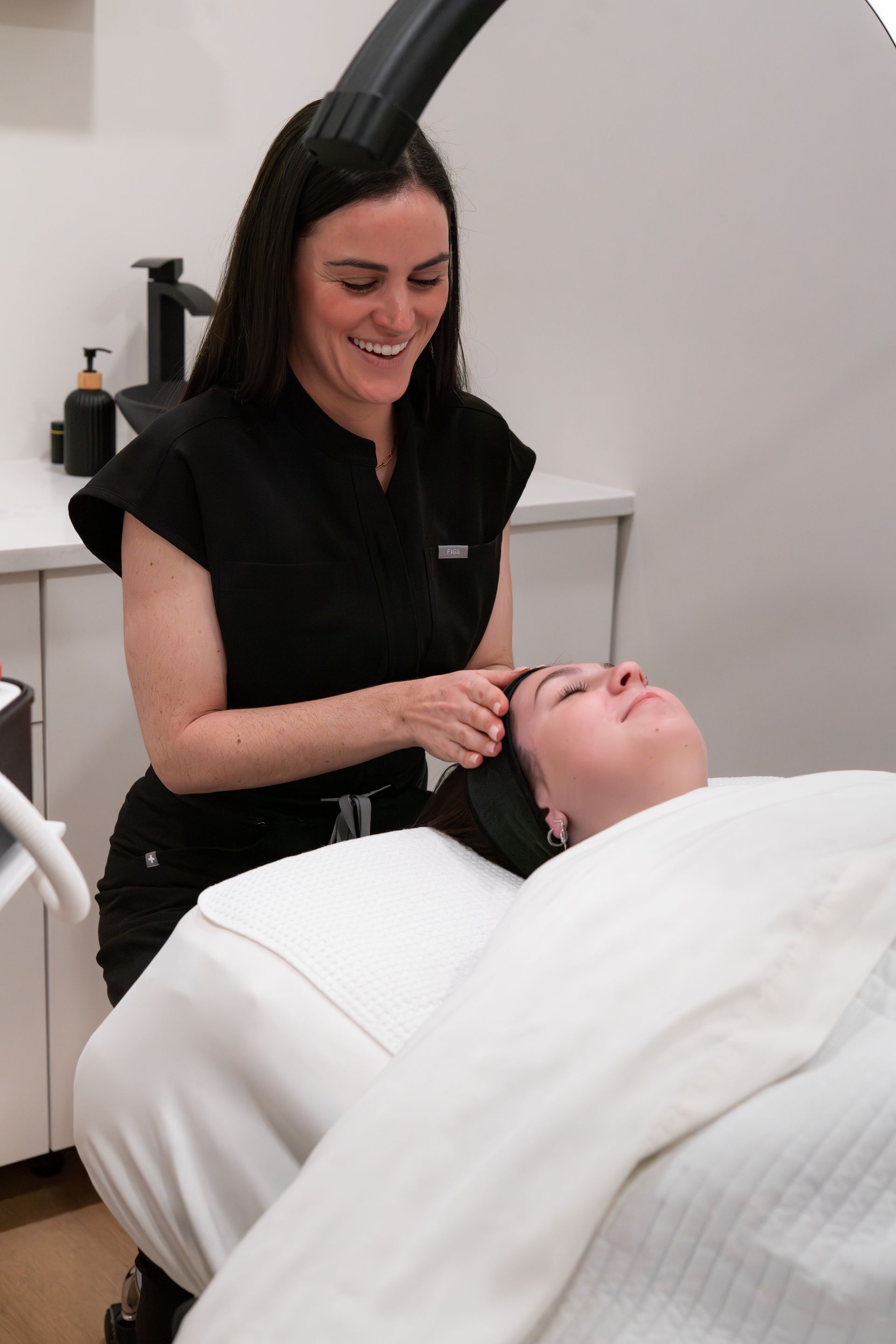 Aesthetic treatment in a spa clinic, practitioner smiling while applying a facial procedure to a reclining client