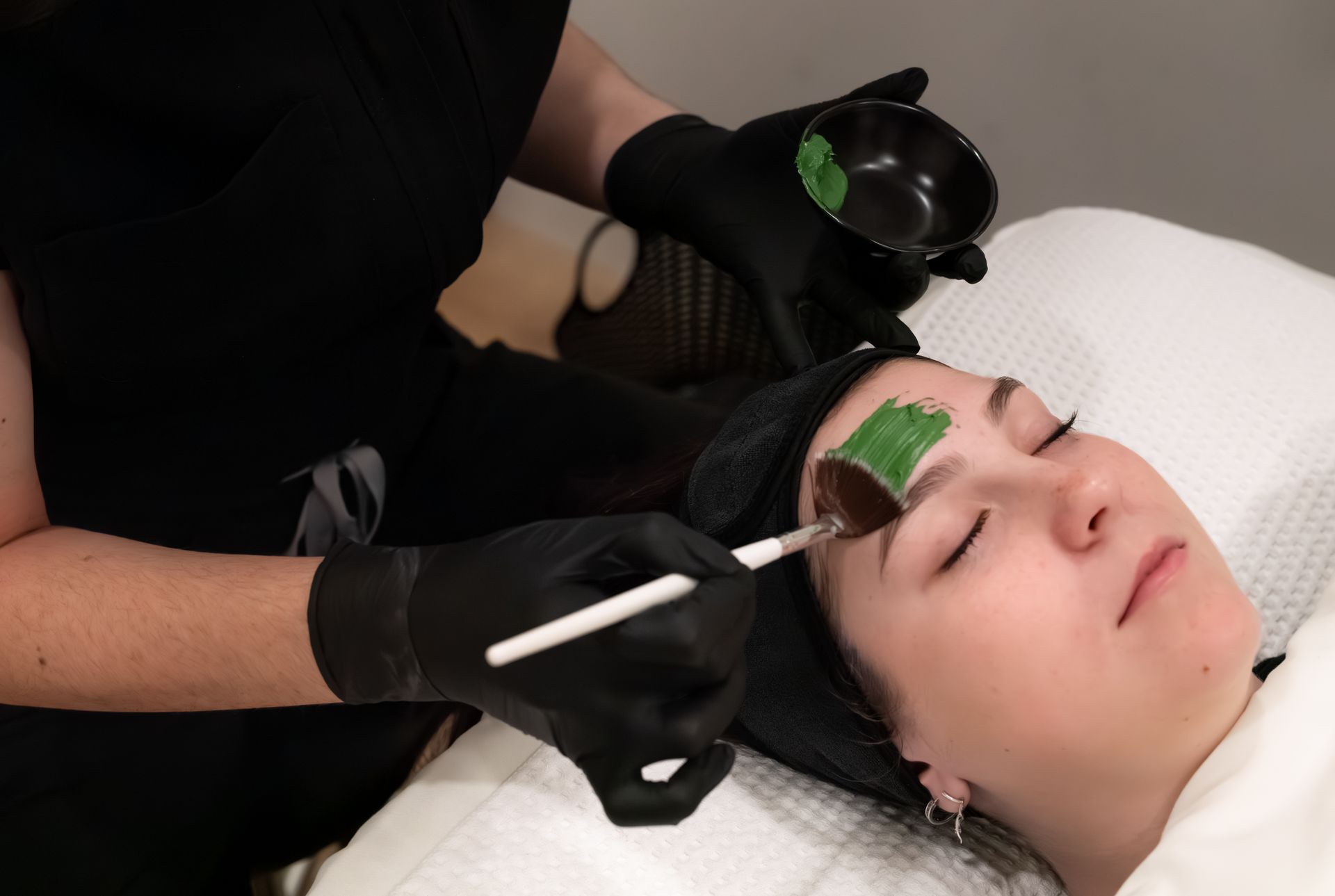 Aesthetic facial treatment with green mask applied to the forehead and a brush in a spa setting