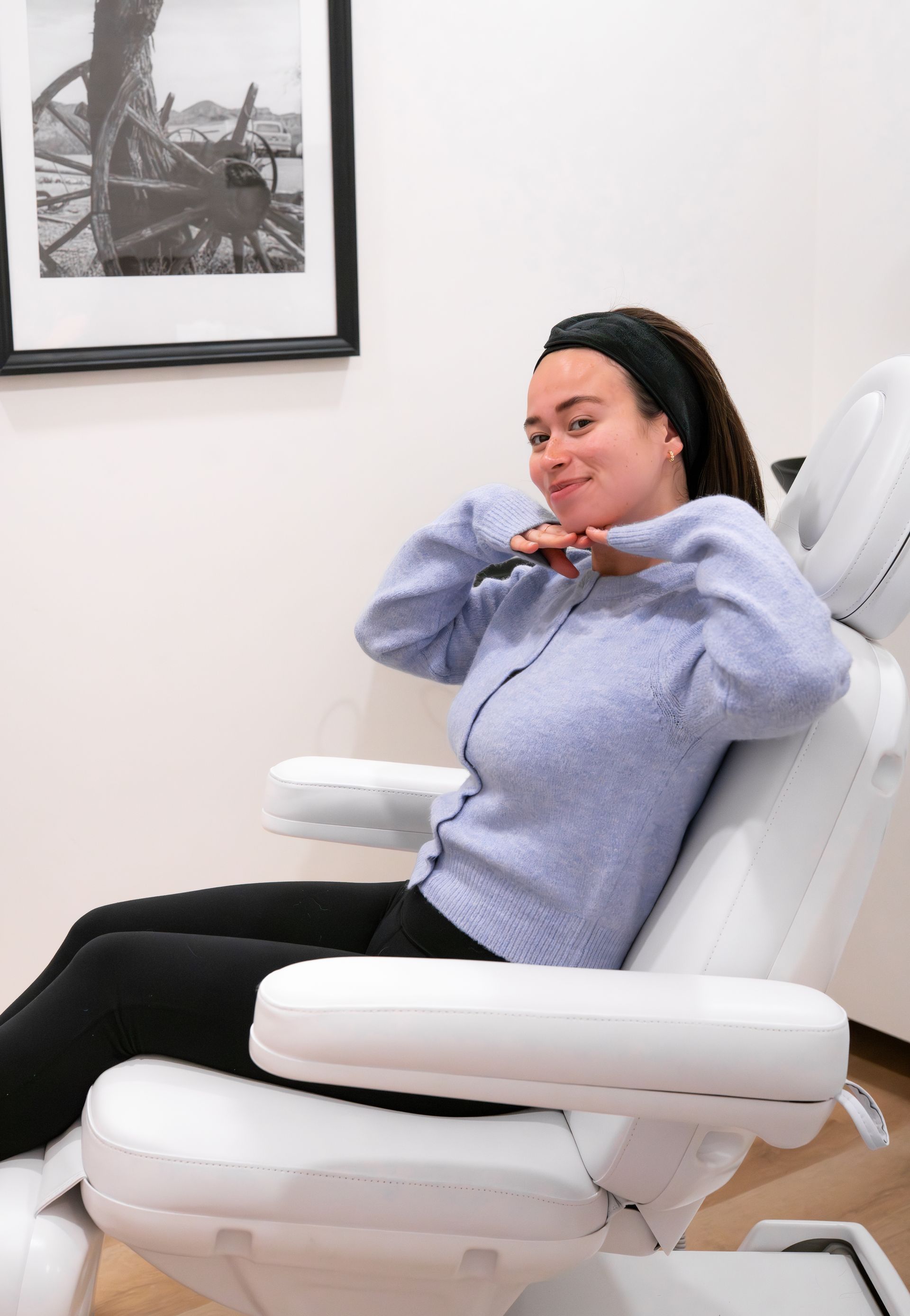Woman reclining in a white salon chair, smiling with hands near her face in a bright room.