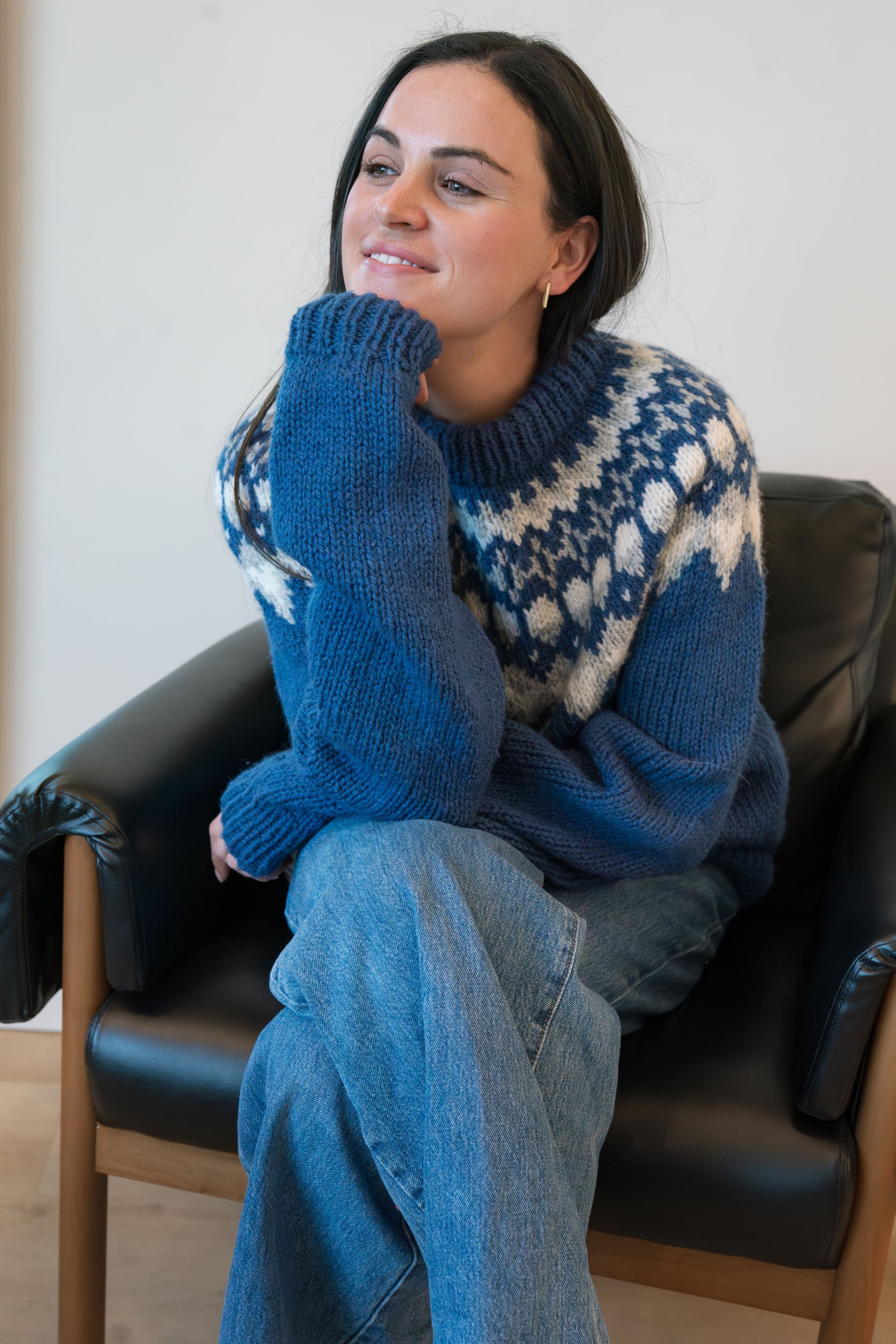 Person seated in a black chair, wearing a blue knit sweater and pants, resting their chin on their hand.