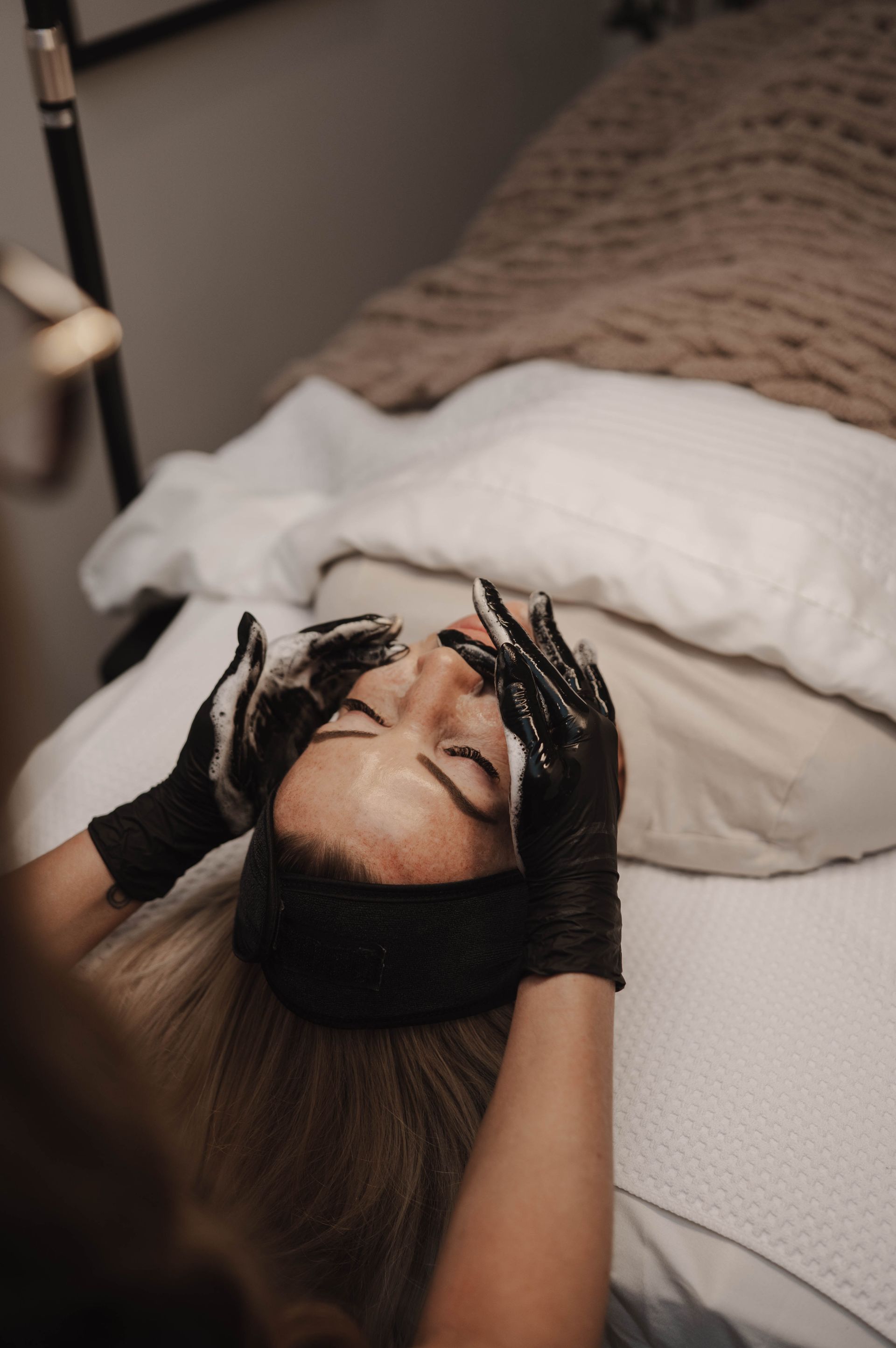 Woman lying on a bed with hands on her face, wearing black gloves and white pillows behind her.