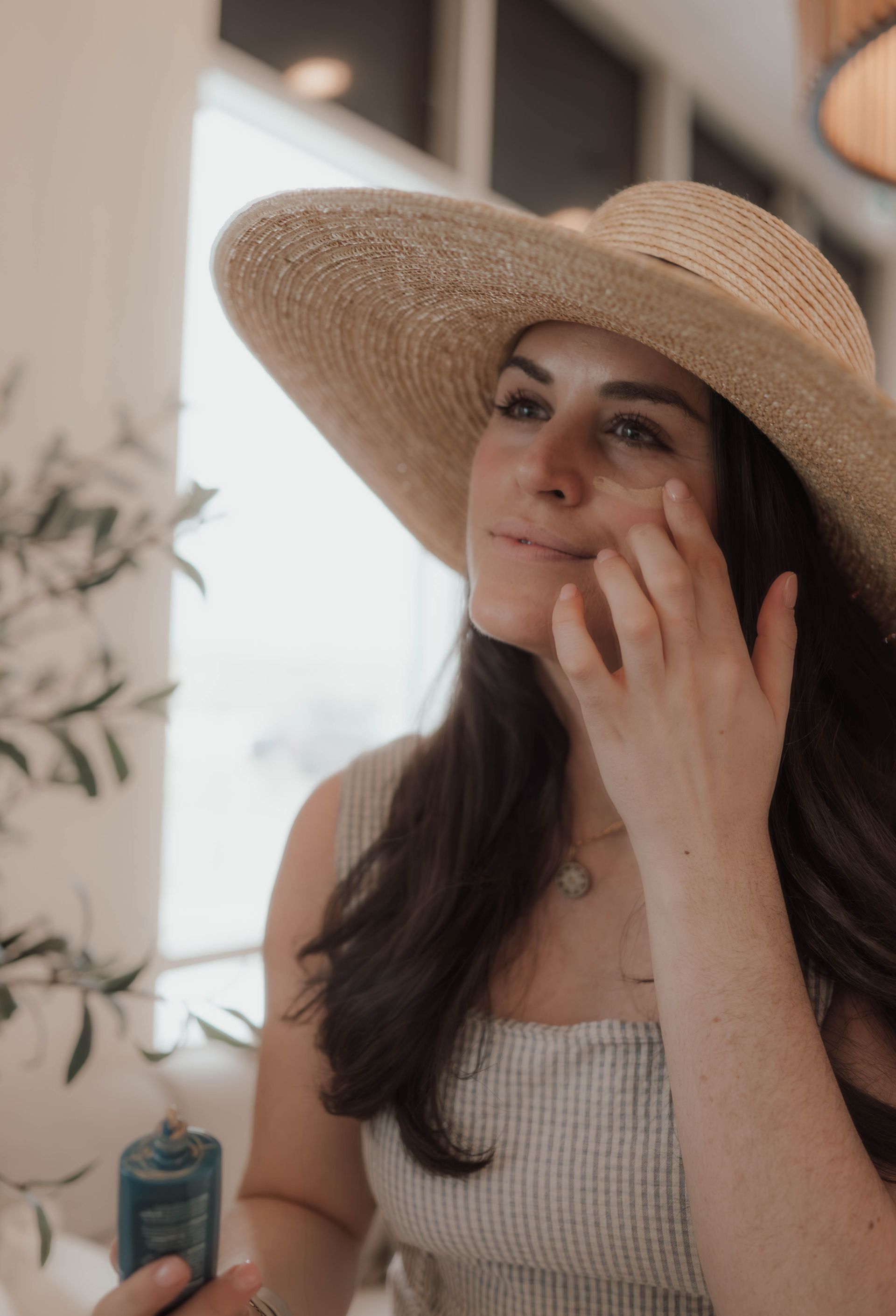 Woman in a straw hat touching her cheek indoors by a bright window