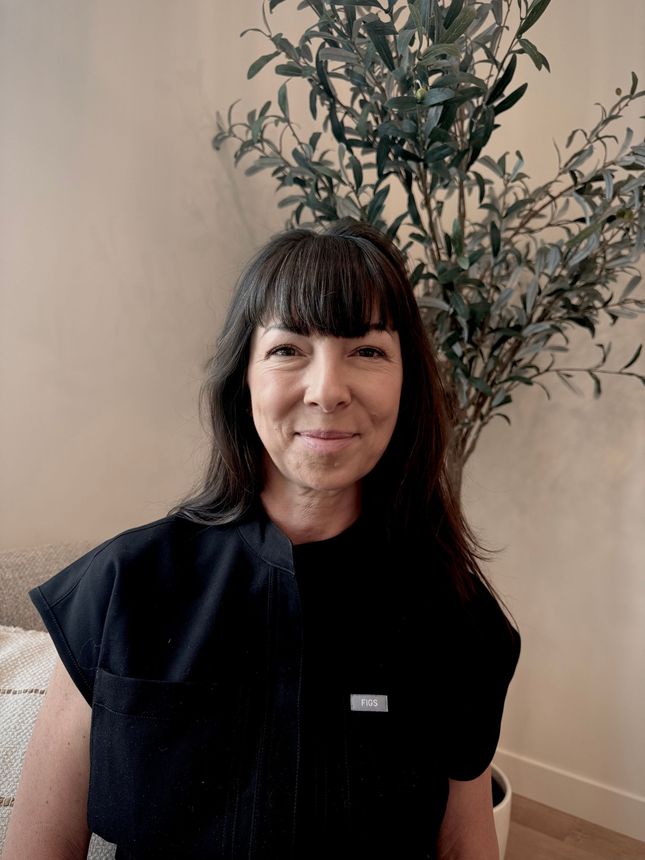 Woman with dark hair standing indoors beside a potted plant, wearing a black top and smiling gently.