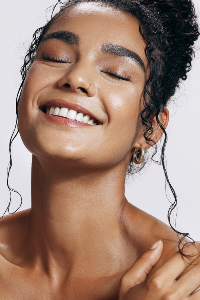 Smiling person with wet skin and closed eyes, touching shoulder against a light background