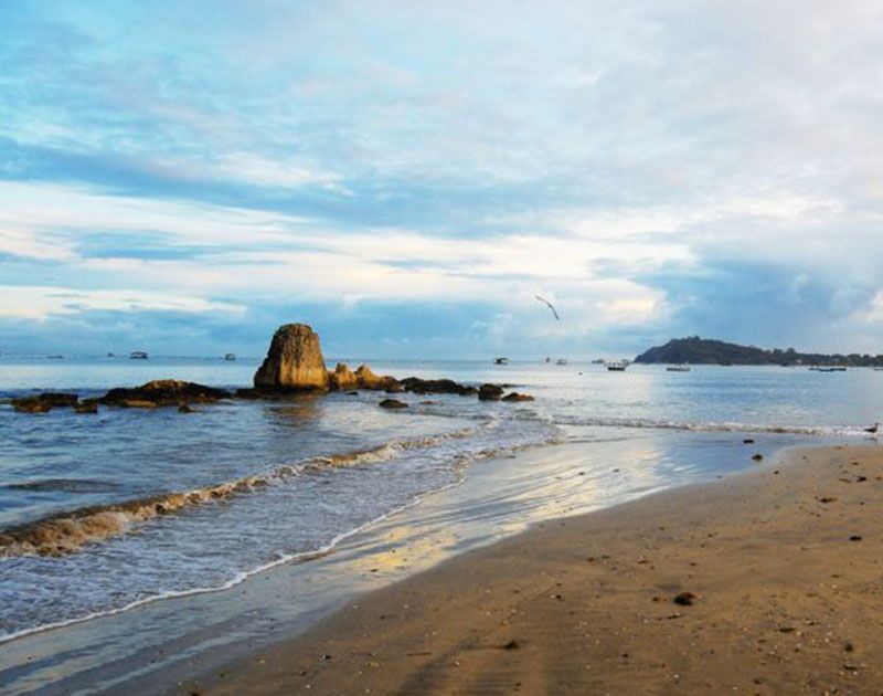 Uma grande formação rochosa emerge do oceano perto de uma praia de areia sob um céu azul e nublado.
