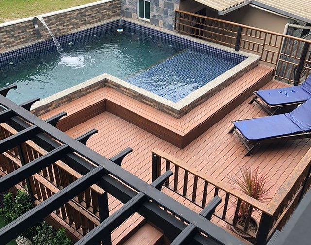 Um terraço elevado ao ar livre com piscina revestida de azulejos, uma fonte e duas espreguiçadeiras azuis sob uma pérgola.