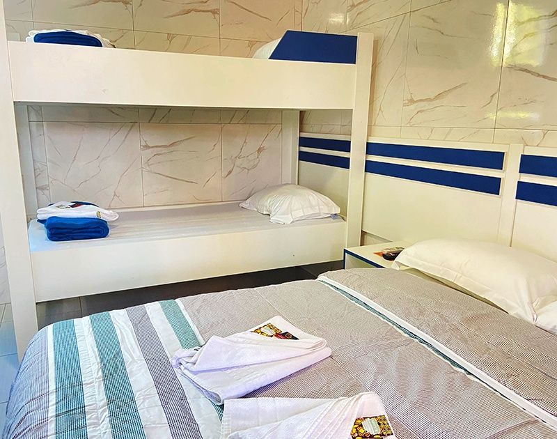 Um quarto com um beliche e uma cama grande, com estrutura branca com detalhes em azul e roupa de cama branca com listras.