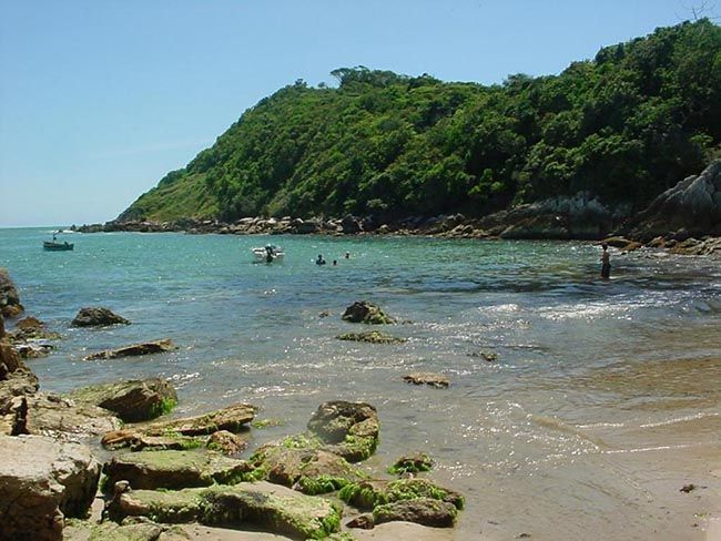 Uma costa rochosa e pitoresca que leva a uma baía de águas turquesa, com uma colina verdejante e exuberante e pessoas nadando ao longe.