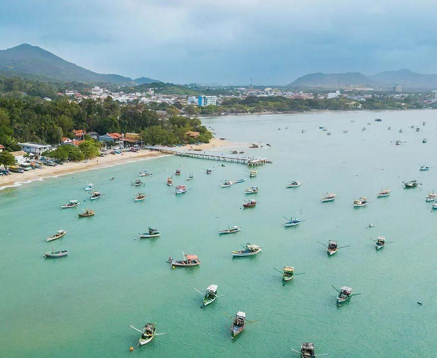 Vista aérea de vários barcos pequenos ancorados em uma baía tranquila de águas turquesa, perto de uma praia arborizada e de uma cidade costeira.
