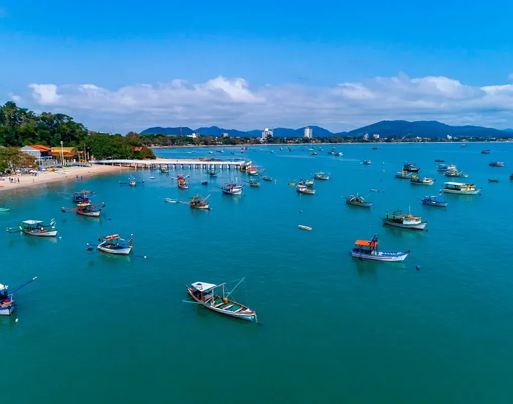 Numerosos barcos de pesca pequenos ancorados em uma baía tranquila de águas turquesa, perto de uma praia de areia e de uma cidade costeira, sob um céu azul.