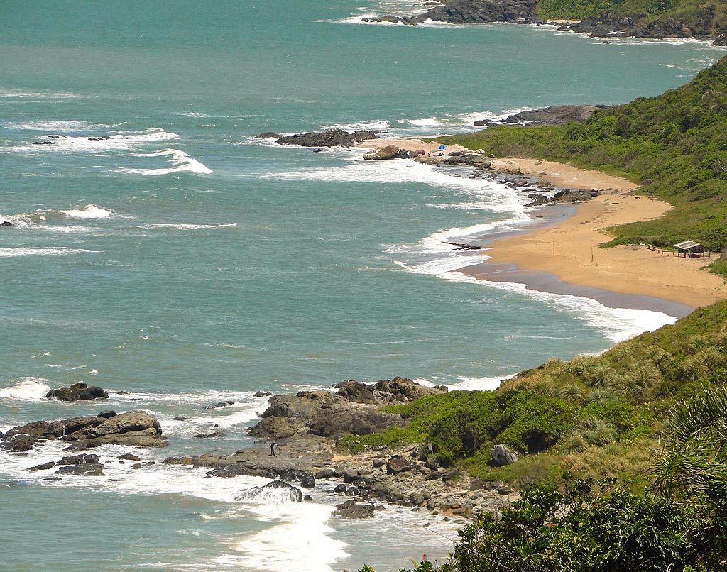 Uma vista elevada de uma praia de areia que encontra águas turquesa, com afloramentos rochosos na costa e colinas verdejantes.