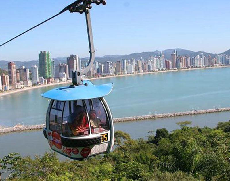 Um teleférico azul, número 35, percorre o céu acima das árvores, com vista para a praia e para o horizonte da cidade em um dia ensolarado.