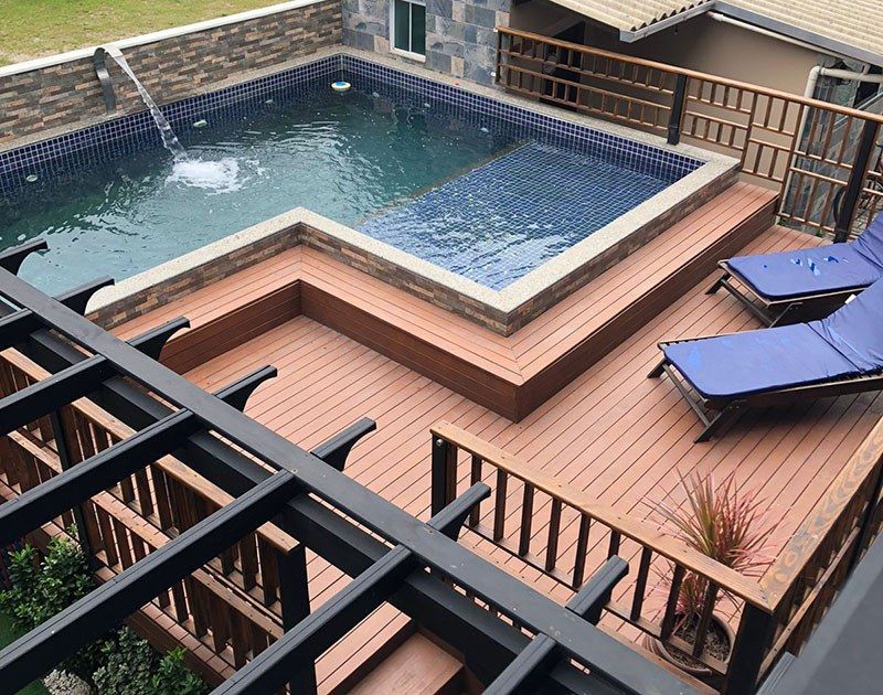 Um deck de madeira com uma piscina elevada com cascata, espreguiçadeiras azuis e uma estrutura de pérgola preta.