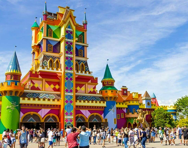 Um castelo colorido e ornamentado, com várias torres e uma grande multidão de pessoas caminhando em frente à entrada.