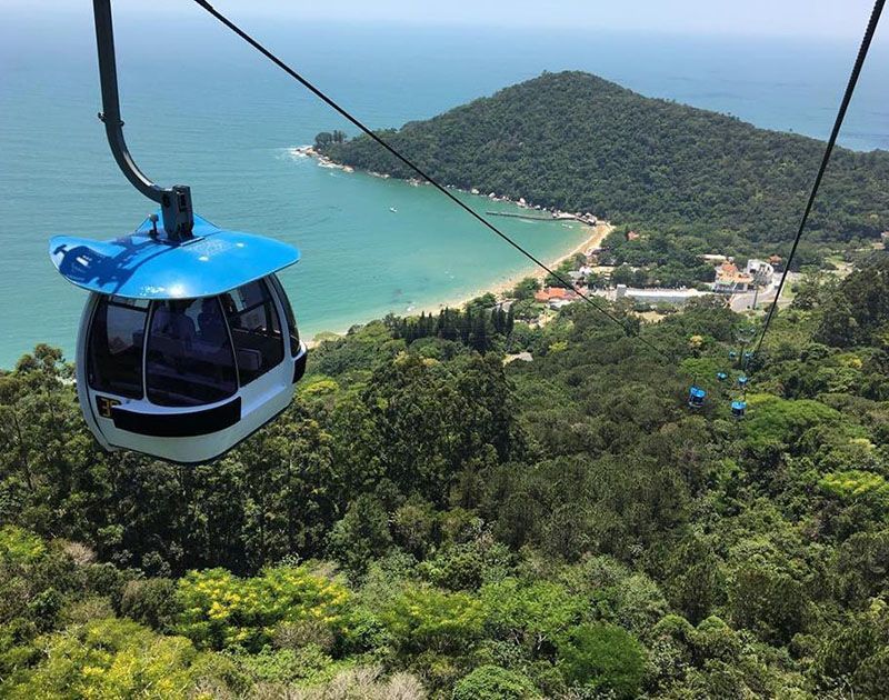 Um teleférico azul e branco desliza sobre uma exuberante floresta verde, com vista para uma praia costeira e o oceano.