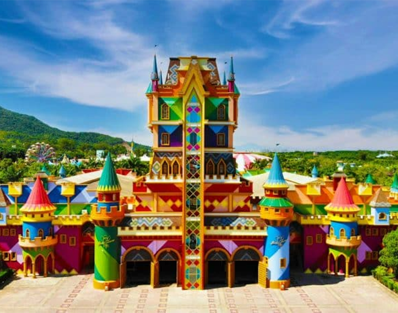 Um castelo colorido e extravagante, com torres altas e padrões geométricos vibrantes, emoldurado por uma paisagem ensolarada e luminosa.