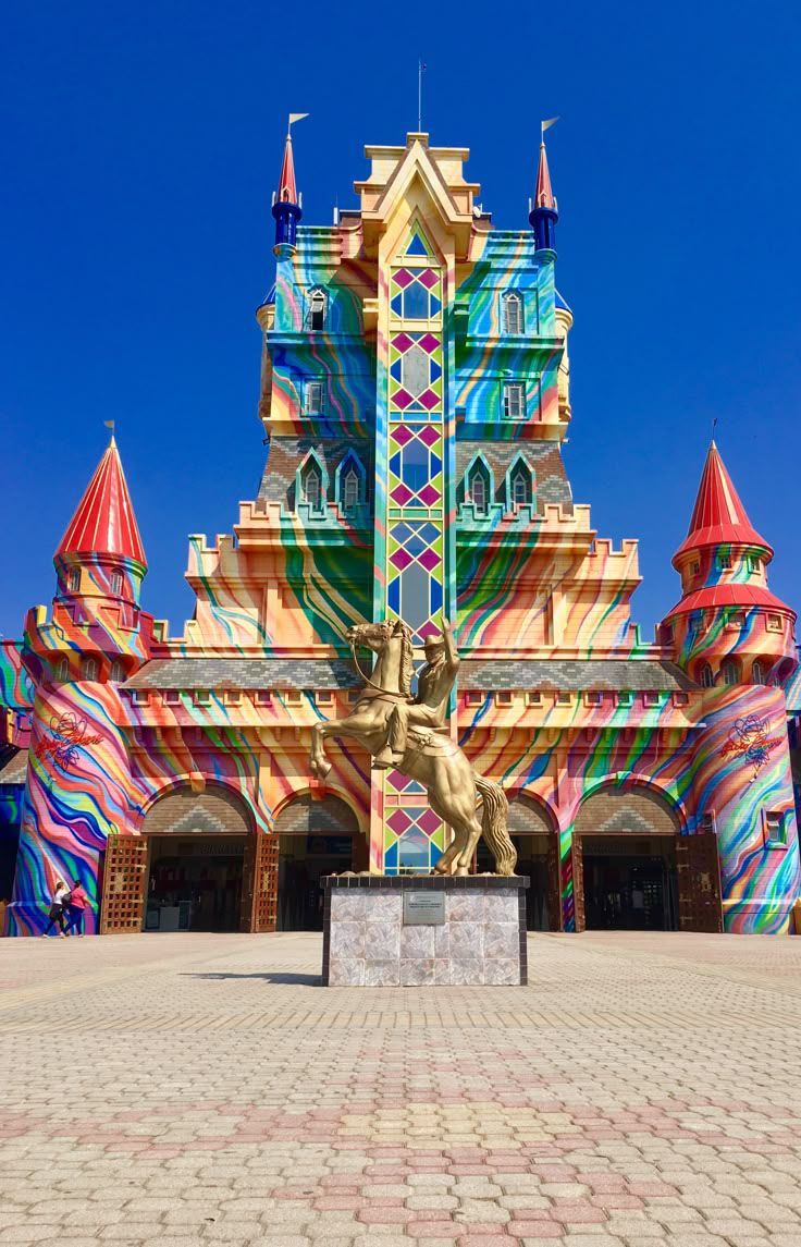 Um castelo multicolorido com padrões vibrantes e uma estátua dourada de um cavalo com cavaleiro em primeiro plano, contra um céu azul claro.