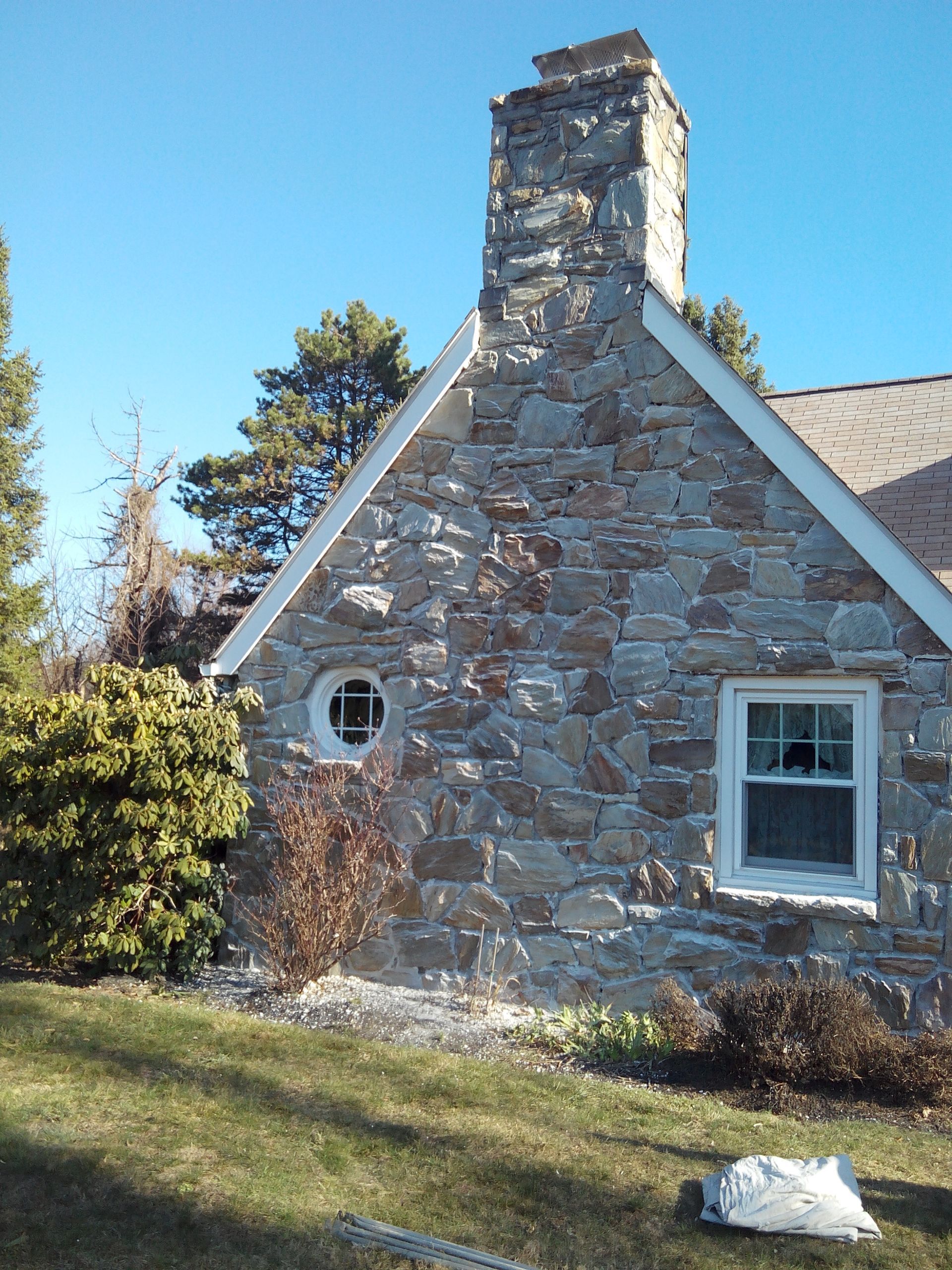 A stone house exterior with a prominent stone chimney, a circular window, and a white-framed rectangular window.
