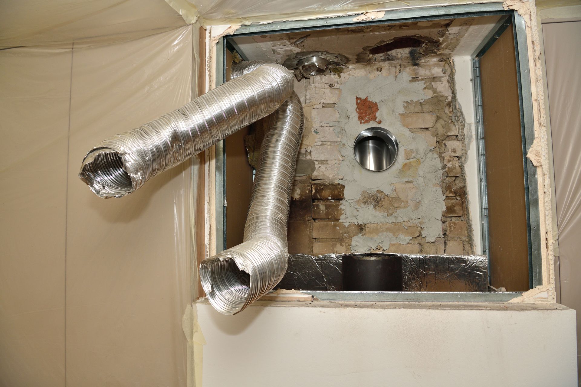 A construction scene showing silver flexible vent hoses extending from an opening in a wall with brickwork behind them.