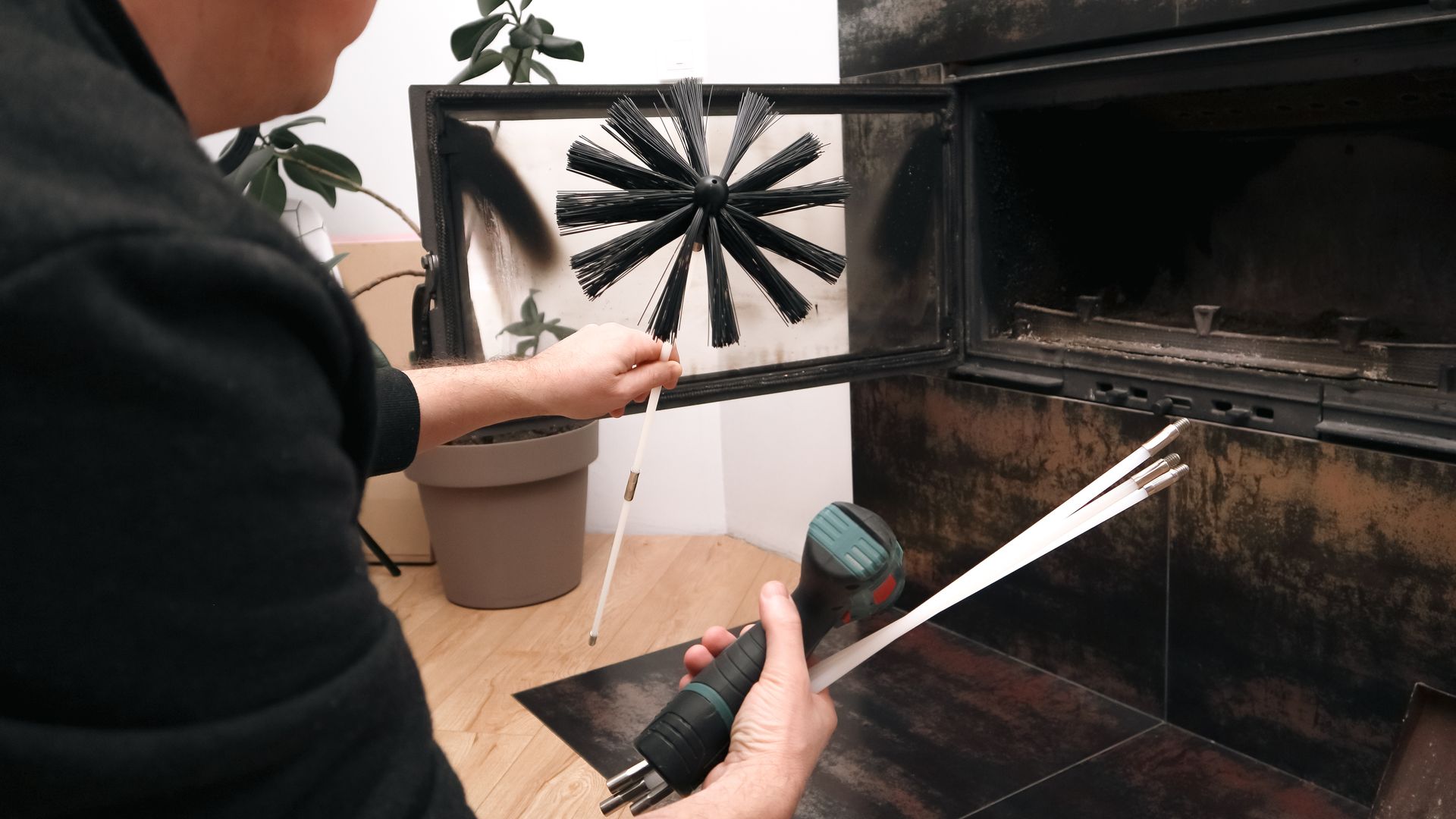 A person holds a chimney brush and an electric drill with extension rods, preparing to clean a fireplace.