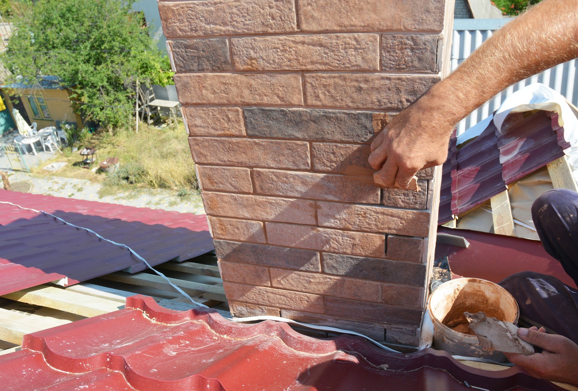 A person uses a trowel to apply mortar between bricks on a chimney structure on a red-tiled roof.