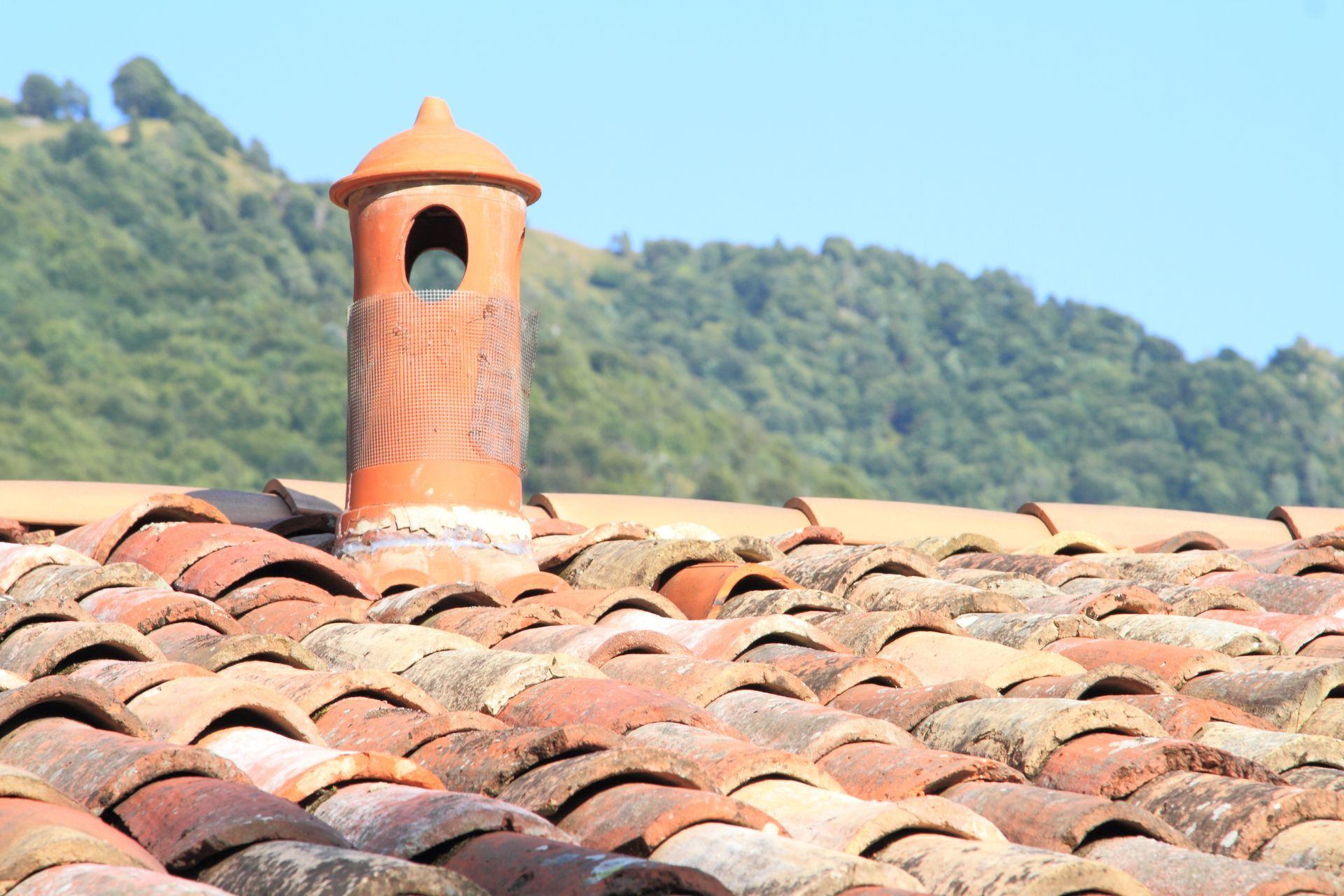 Terracotta roof tiles with a small orange chimney against a green hillside and blue sky