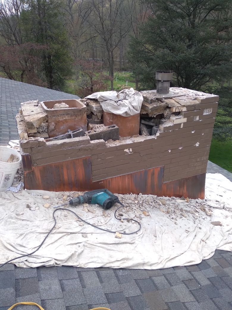 A person in a gray shirt and shorts builds a brick chimney on a corrugated roof.