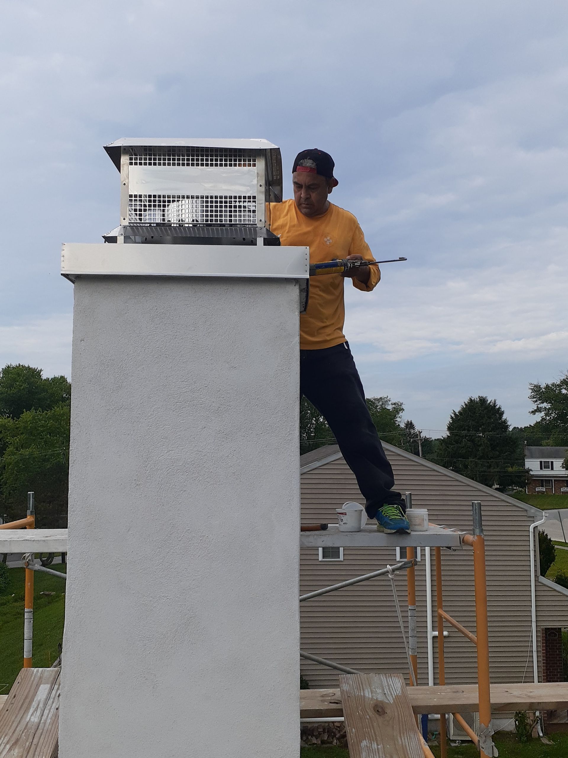 A worker in a yellow shirt stands on scaffolding, installing a metal chimney cap on a stone chimney.
