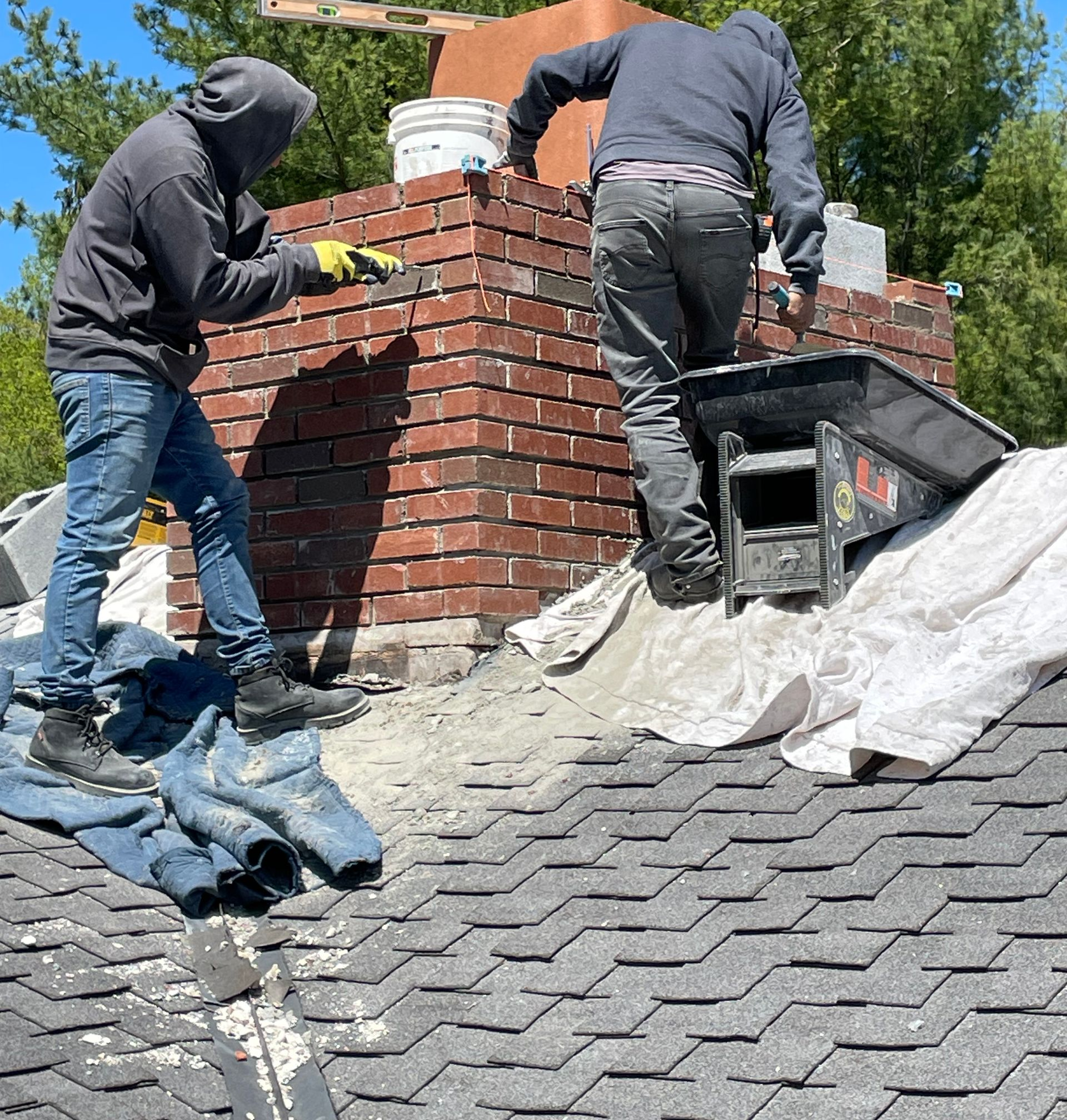 A person repairs a weathered brick chimney on a sloped roof surrounded by lush green trees.