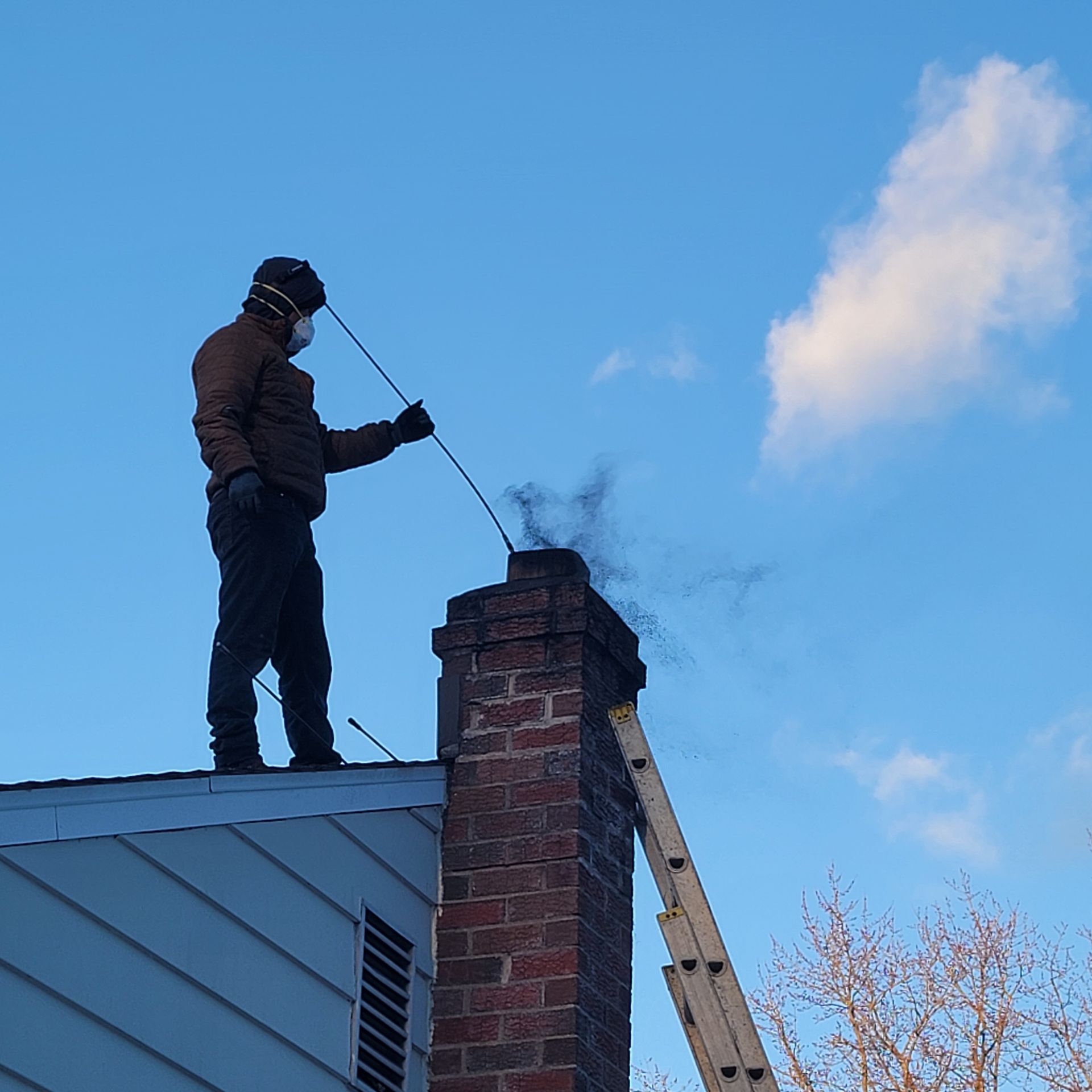 A worker in dark clothing stands on a tiled roof, positioned next to a chimney while holding tools.