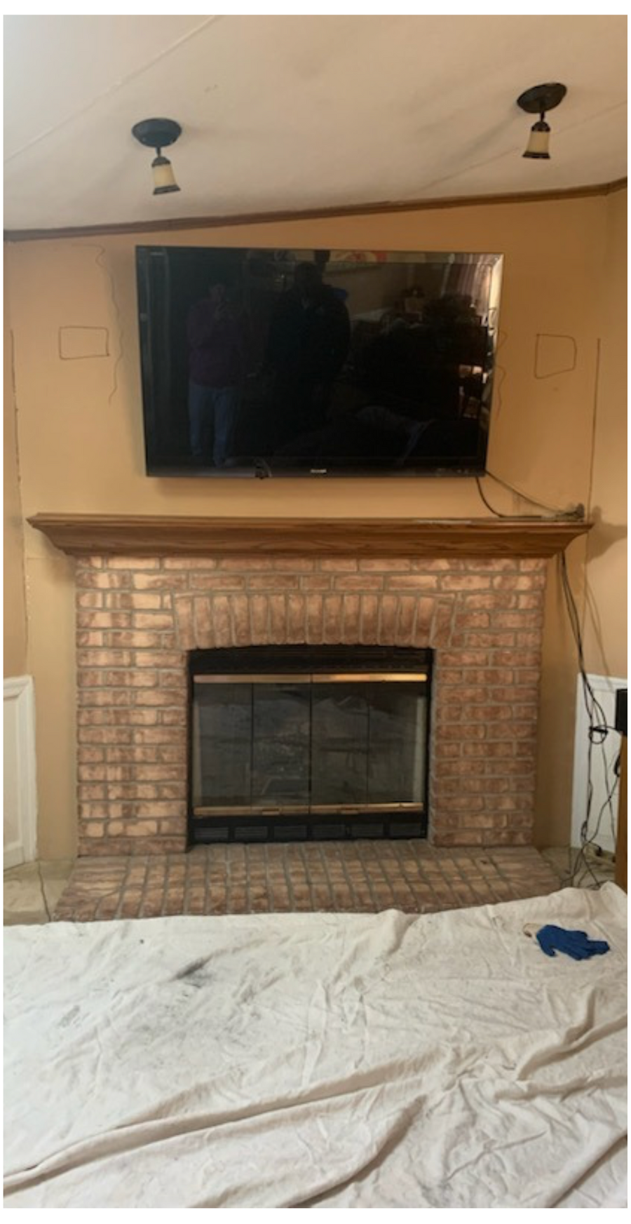 Living room fireplace with large wall-mounted TV, beige walls, white drop cloth, and exposed cords