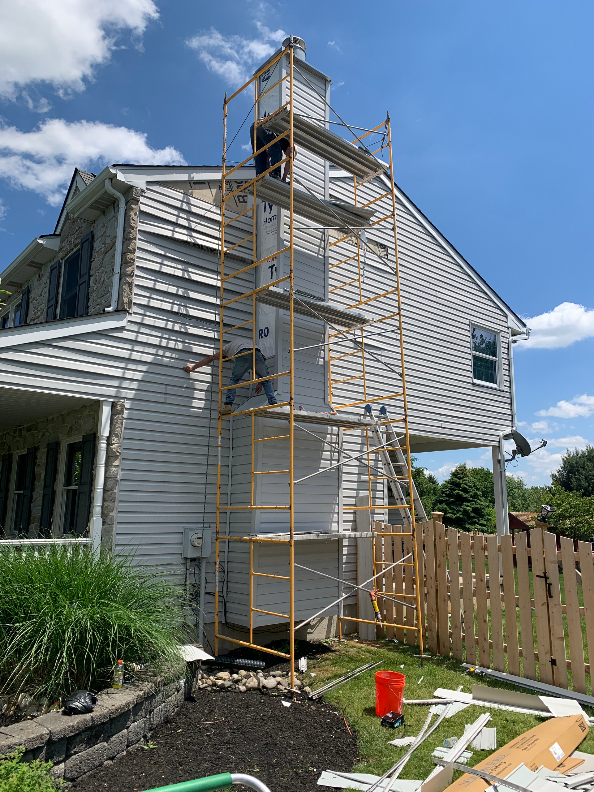 A person works on scaffolding around a white siding chimney on the side of a two-story house under a blue sky.