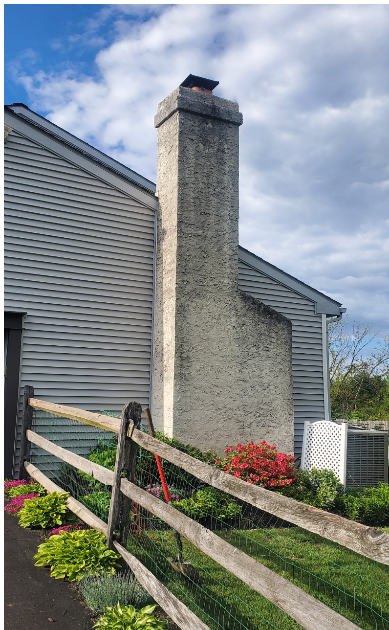 Weathered stone chimney beside gray siding and a wooden fence under a blue sky