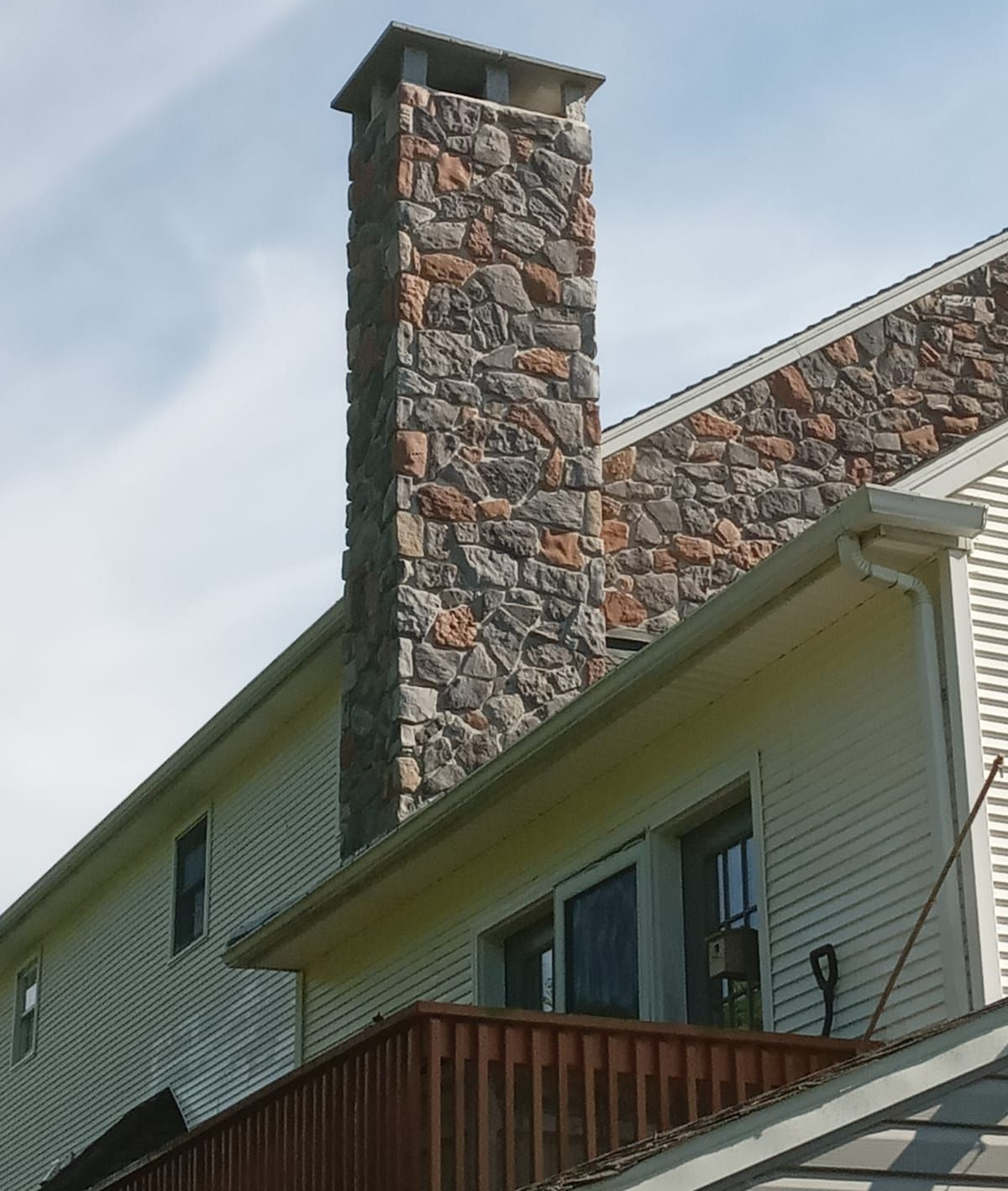 A person in dark clothing stands on the snowy roof of a two-story house next to a chimney during the day.