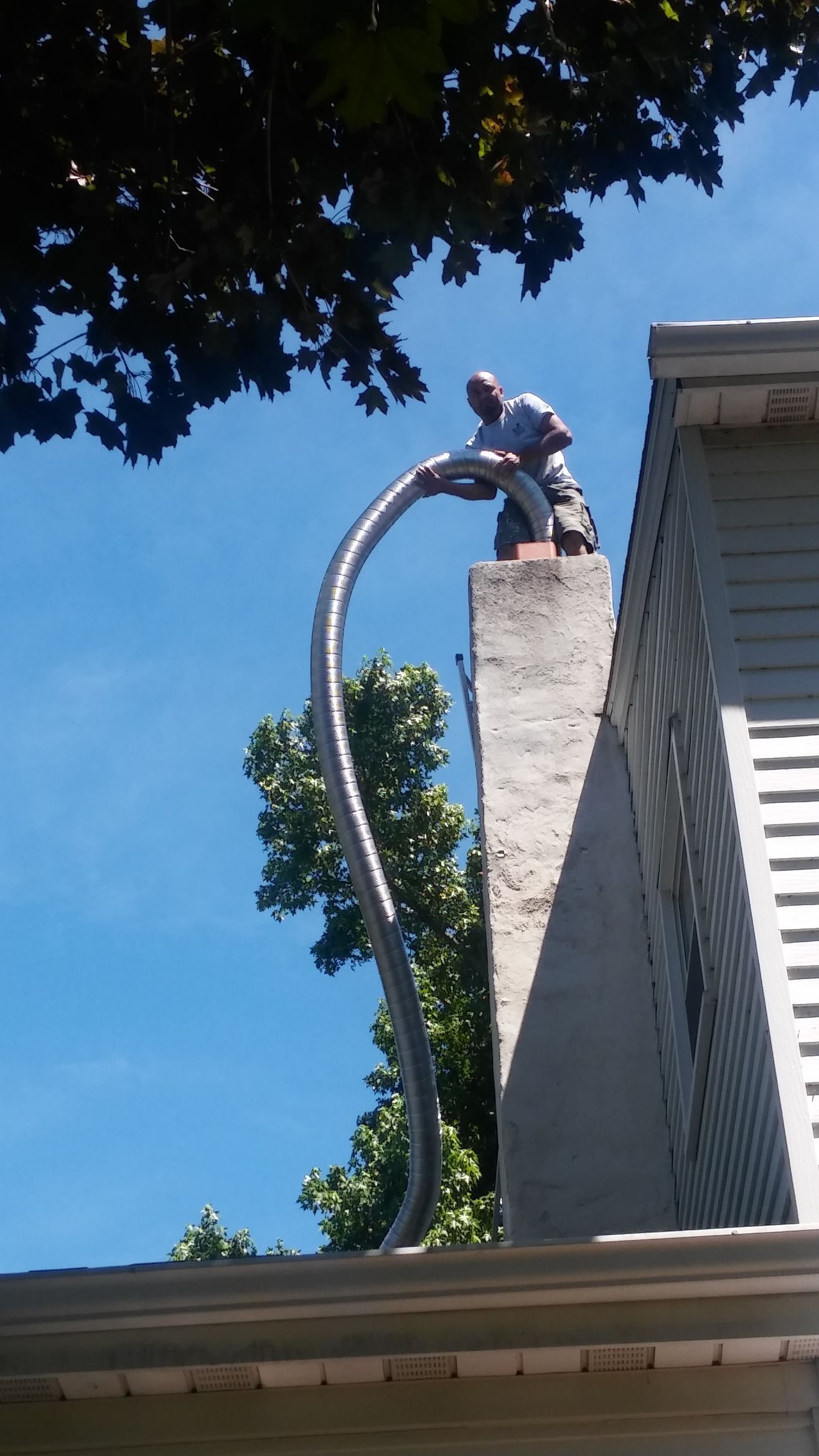 A person standing on a sloped shingled roof, leaning over a tall brick chimney with white bags stacked at its base.