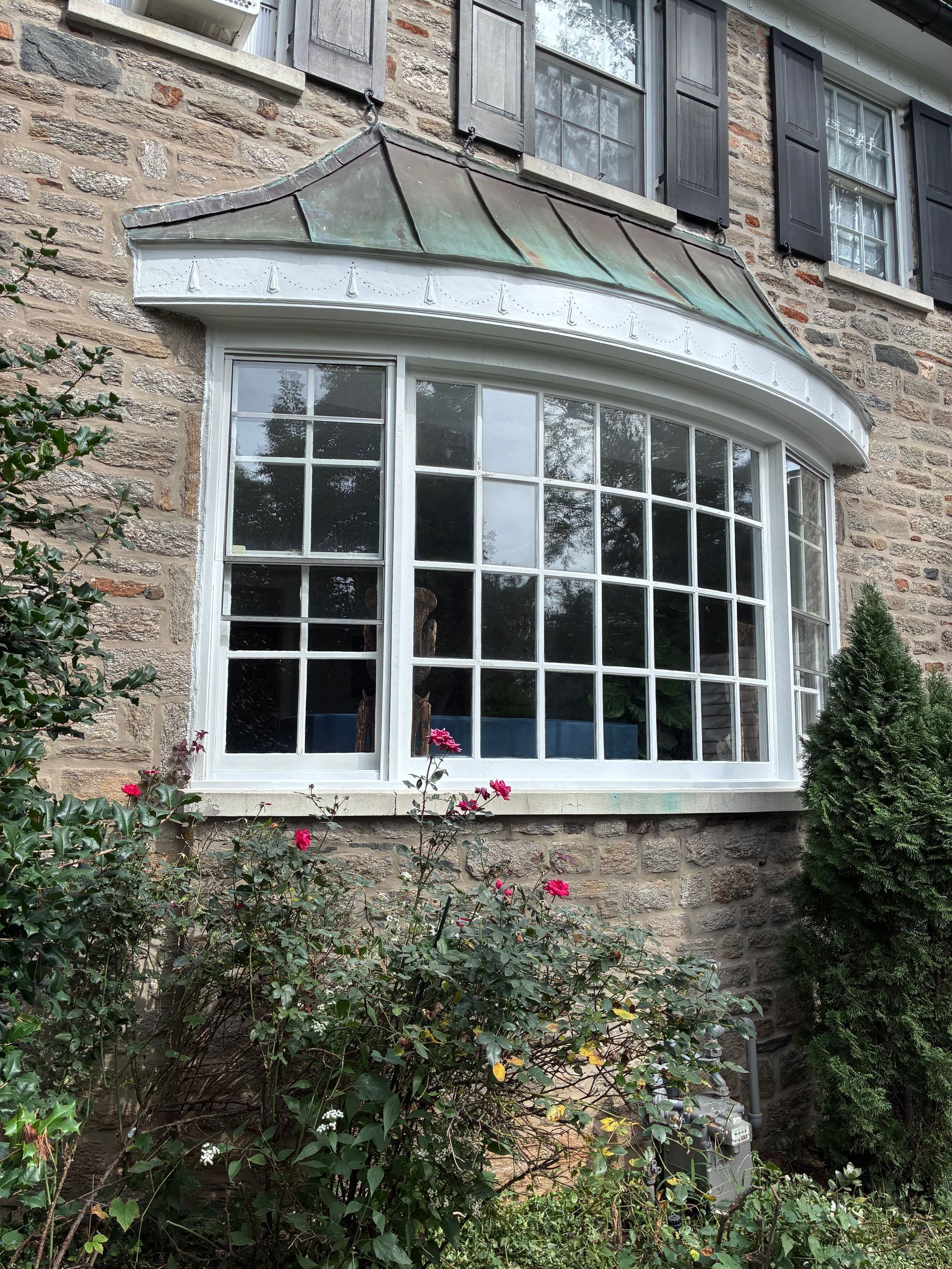 Bay window with multi-paned glass on a brick building, under a copper-colored roof. Bushes and small rose flowers in front.