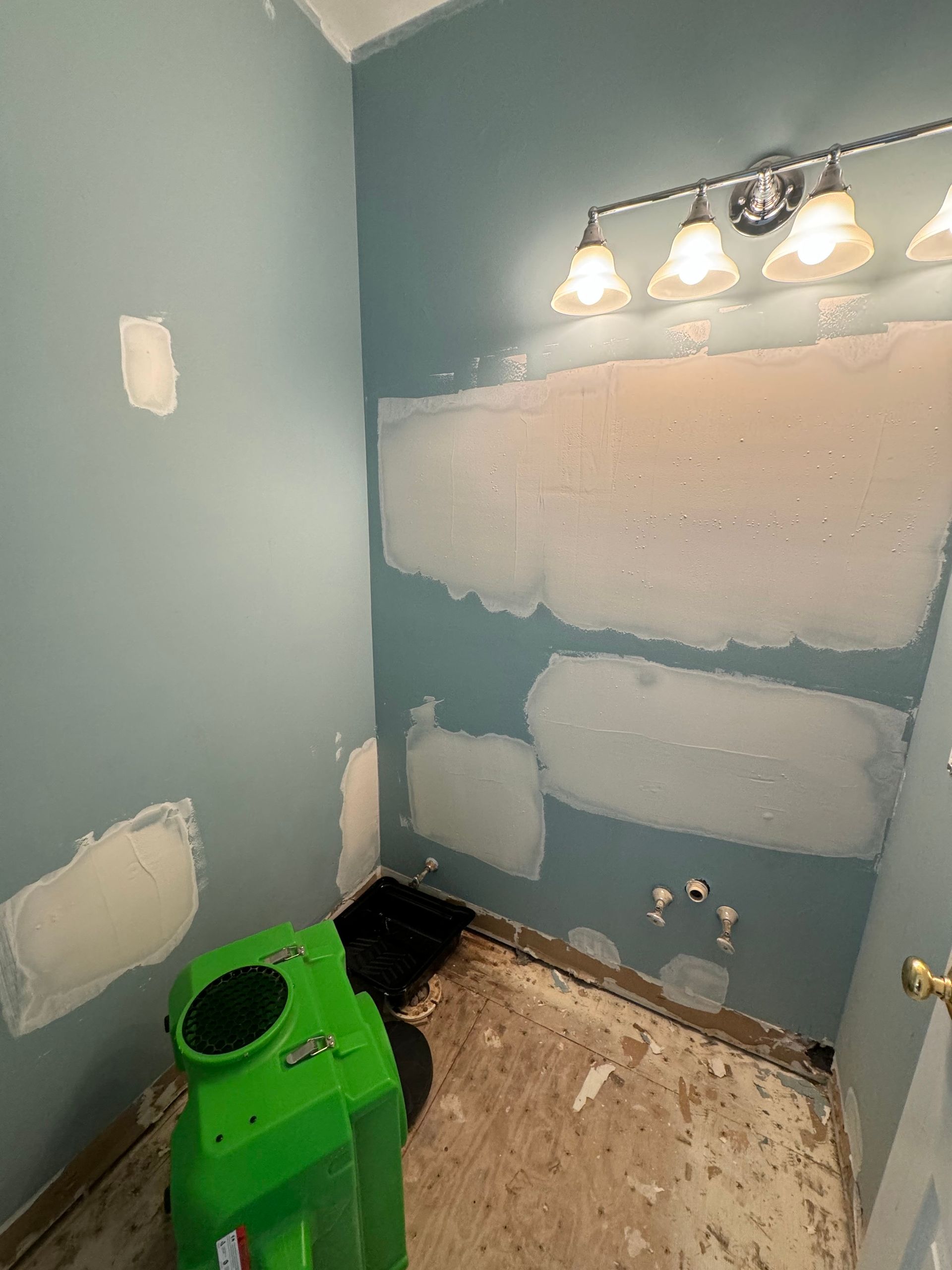 Bathroom renovation in progress; teal walls with drywall patches, green dehumidifier, exposed pipes, and a light fixture.